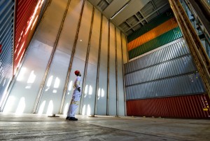 Stacks of shipping containers inside the hold of a cargo ship with a cargo ship worker inspecting the containers