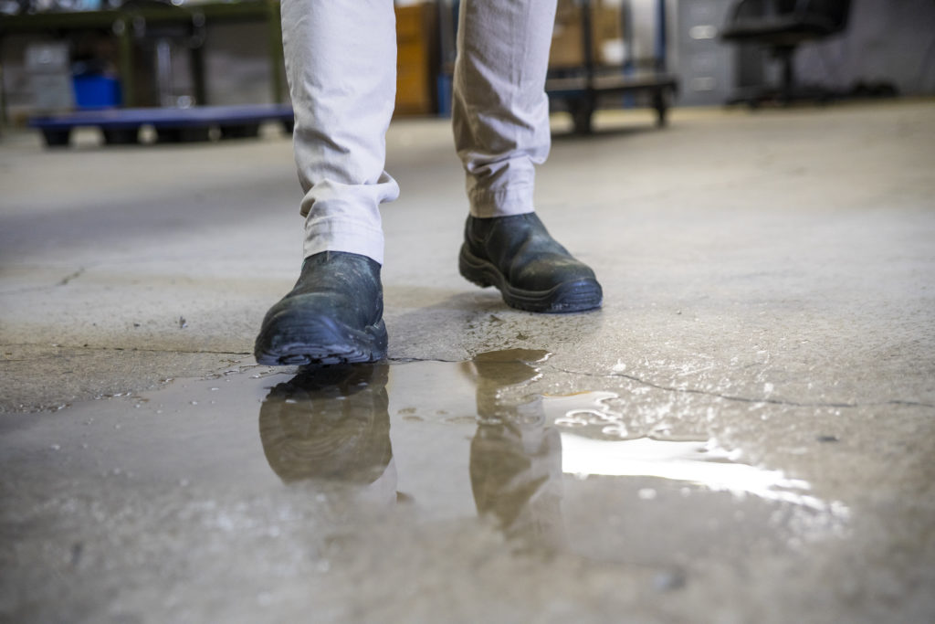 A male worker wearing work boots in a warehouse walking into a liquid spill on the floor.